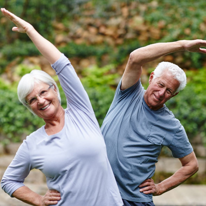 Two elderly people stretching outdoors with greenery in the background