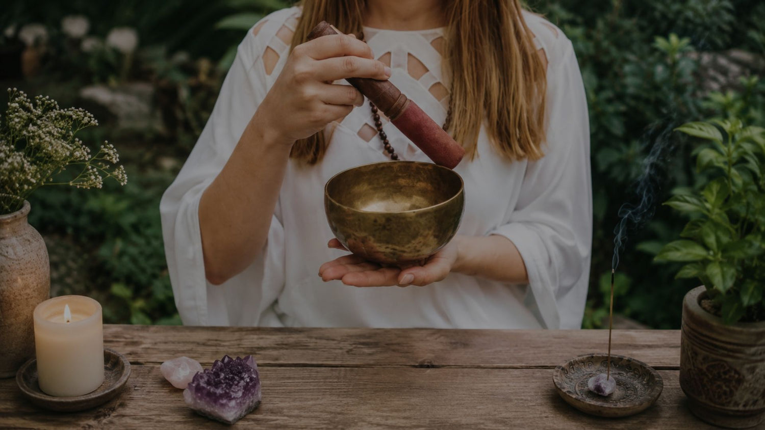 Person holding a singing bowl with incense and candles on a wooden table outdoors.