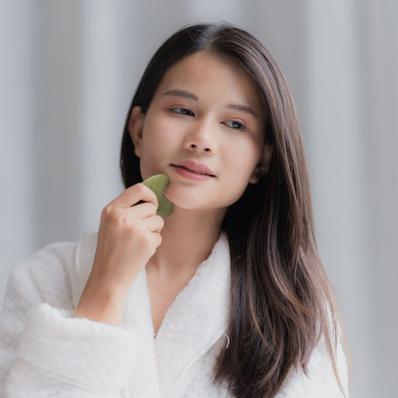 Woman in a white robe holding a green facial tool against her face.