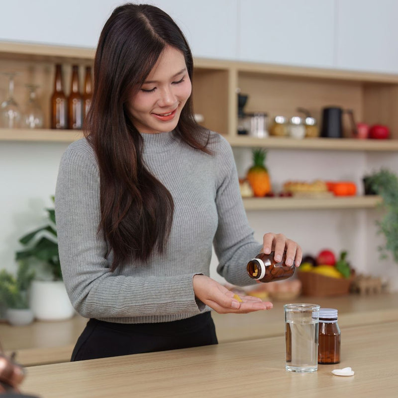 Woman in a kitchen pouring a pill into her hand with a glass of water on the counter.